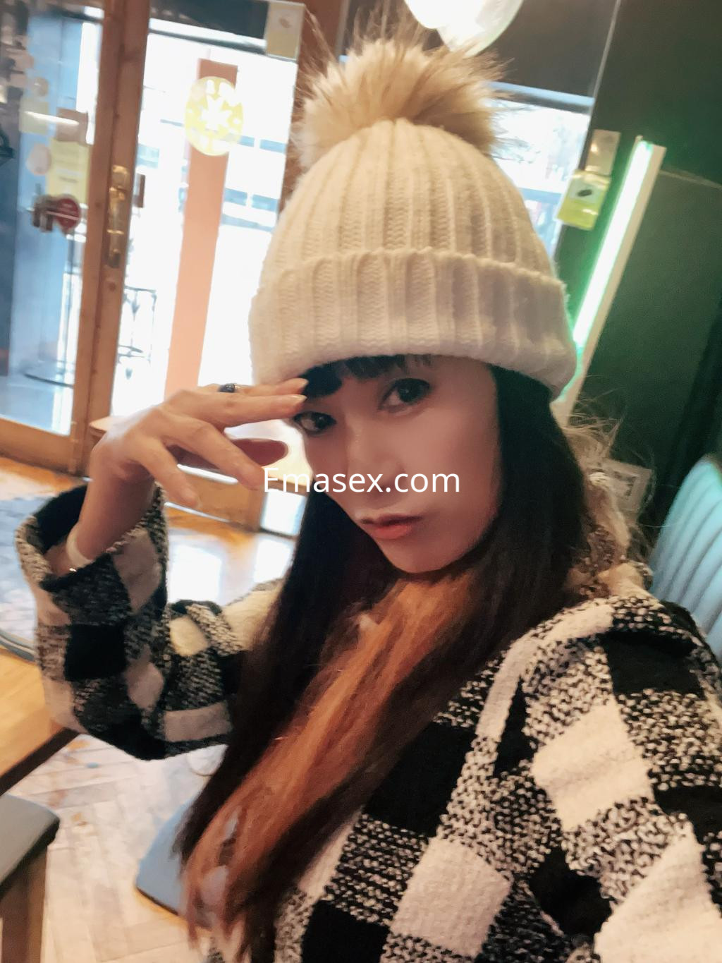 Escort en Burgos con gorro blanco de pompón y abrigo a cuadros, posando en un ambiente acogedor.