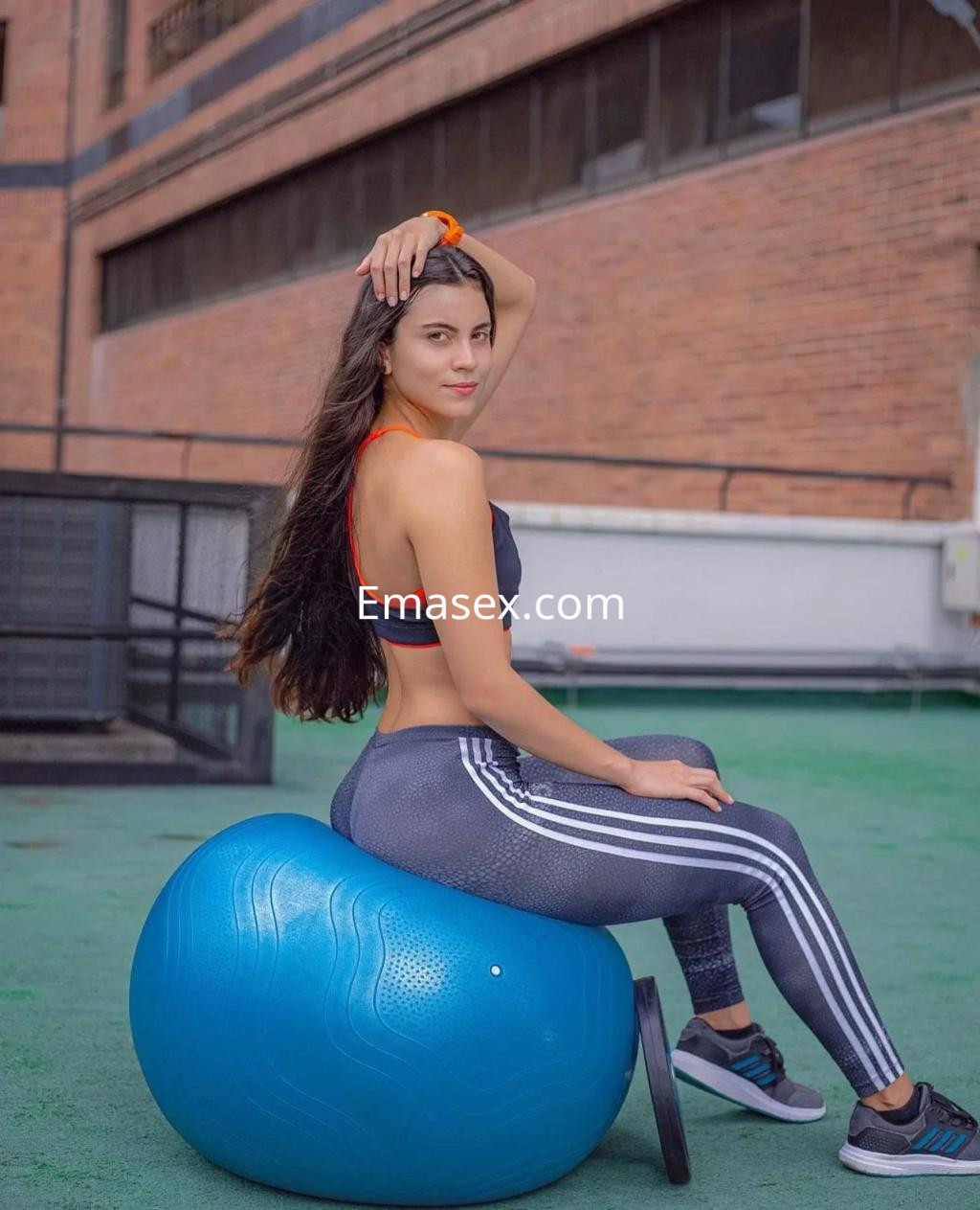 Escort en Sevilla sentada sobre una pelota de ejercicio, luciendo un top deportivo rojo y leggings con rayas.