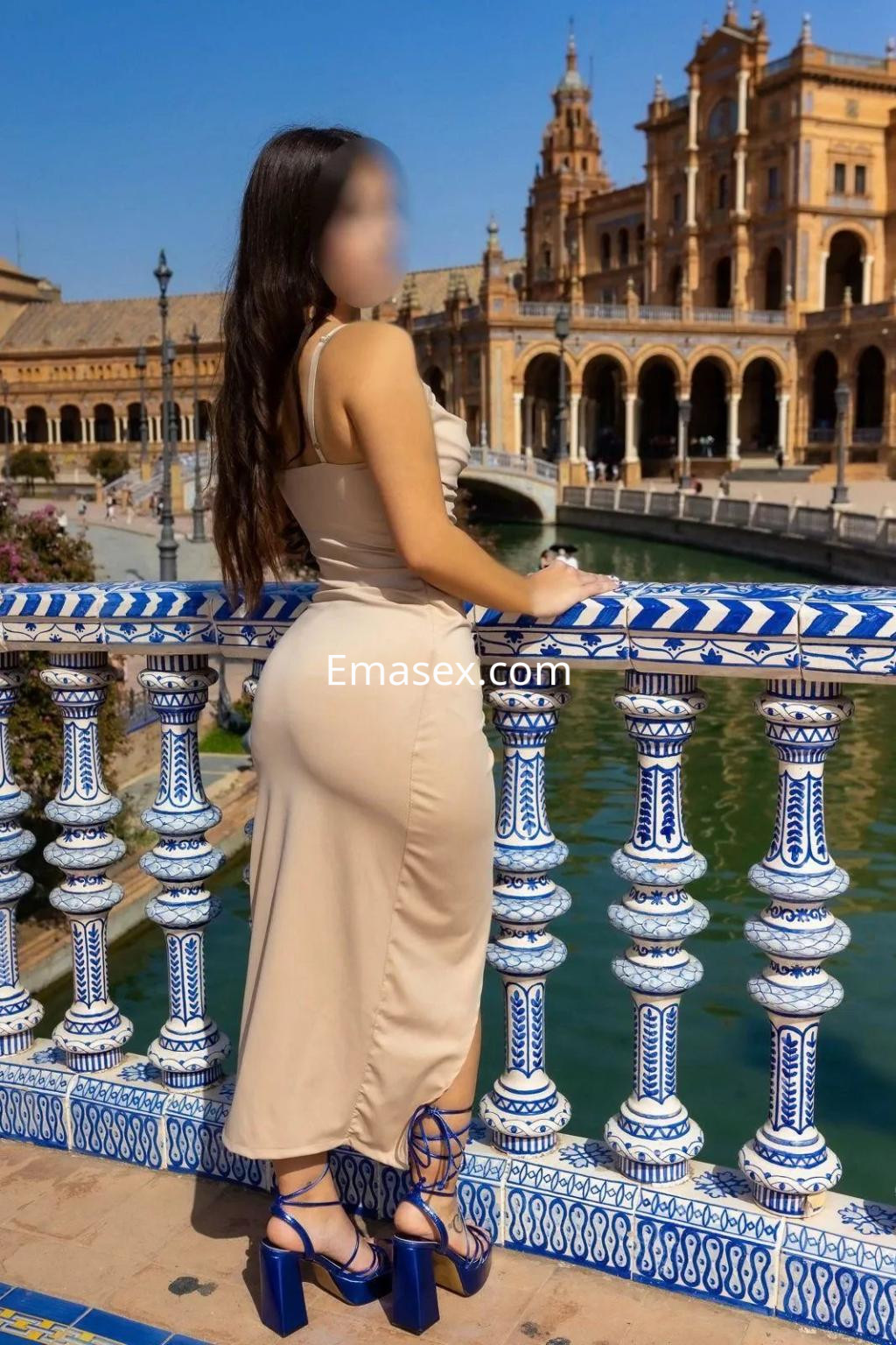 Puta posando de espaldas junto al agua en Sevilla, con un vestido ajustado de color beige y tacones azules.