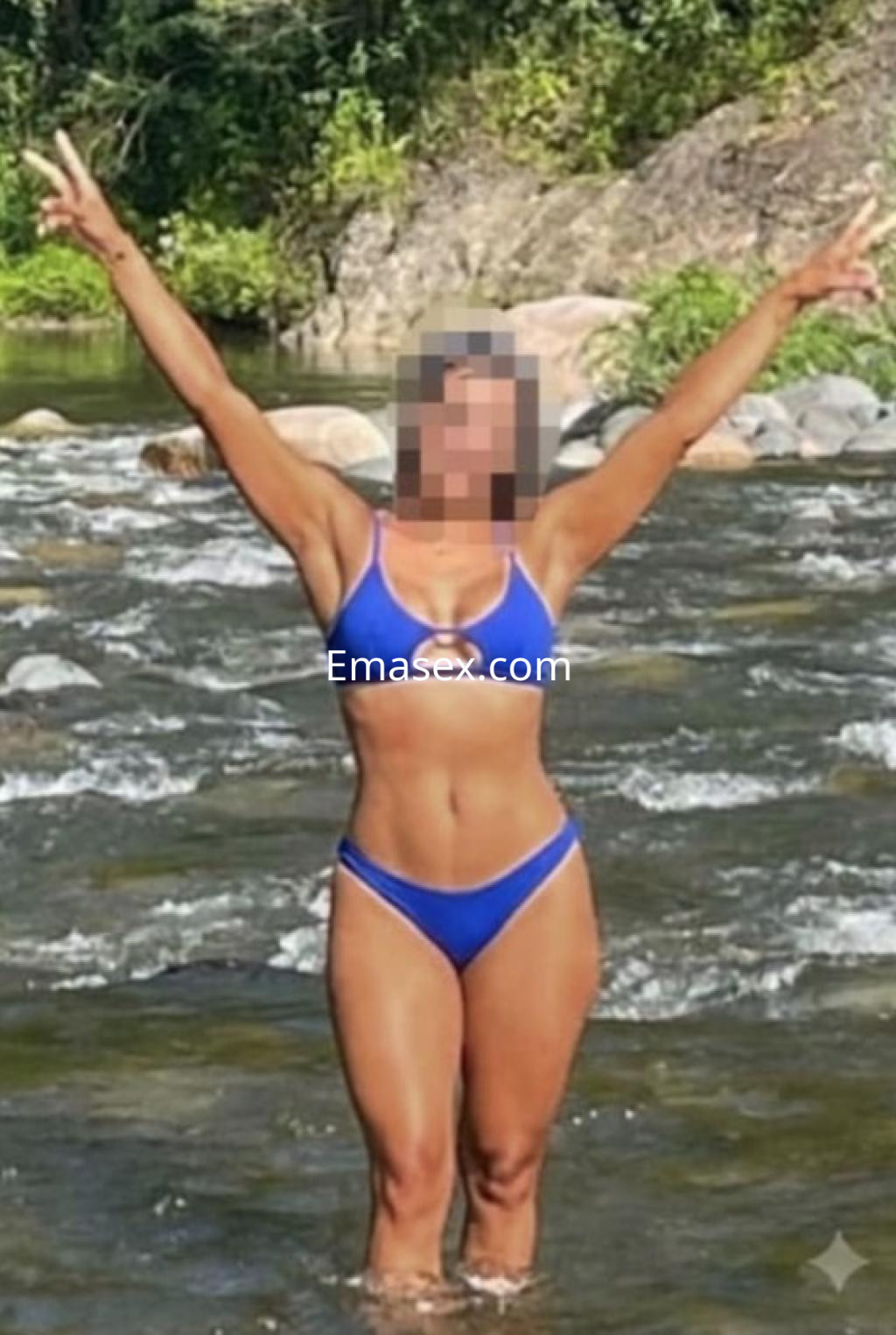 Escort en Coruña de pie en un río, levantando los brazos, luciendo un bikini azul y con el cabello suelto.