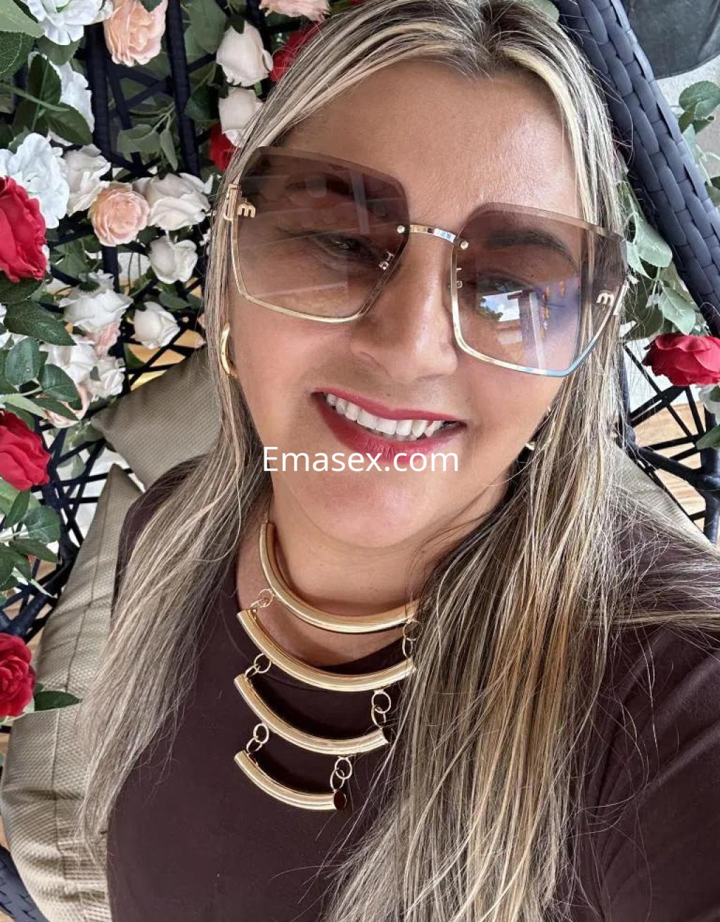 La puta en Badajoz lleva gafas grandes y un collar dorado, sonriendo entre flores decorativas.