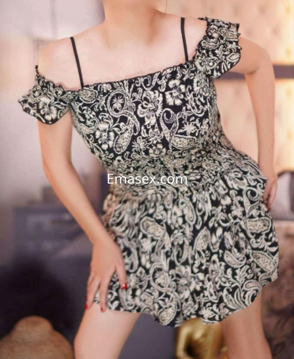 Puta en Madrid con vestido negro con estampado floral, posando de forma seductora en un entorno acogedor.