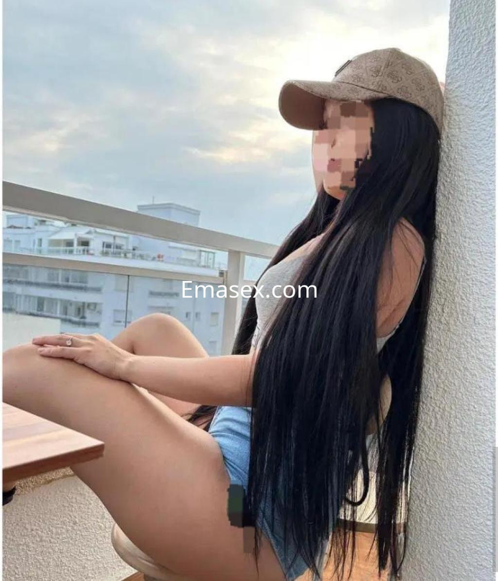 La puta en Albacete luce un top gris y shorts de mezclilla, sentada en un balcón con cabello largo y oscuro.