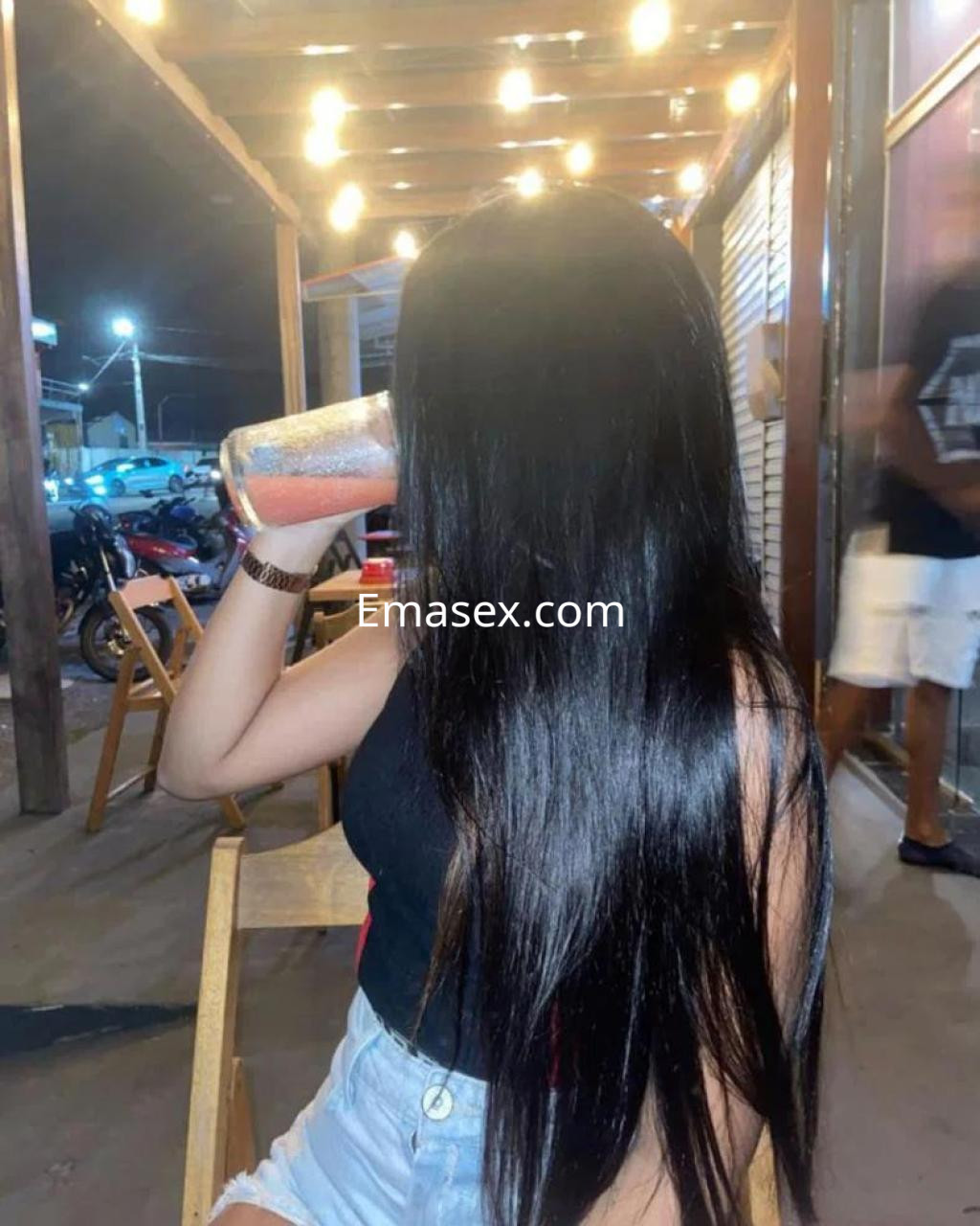 Escort en Cáceres con cabello largo y oscuro, vistiendo camiseta negra y shorts, sosteniendo un vaso en una terraza.