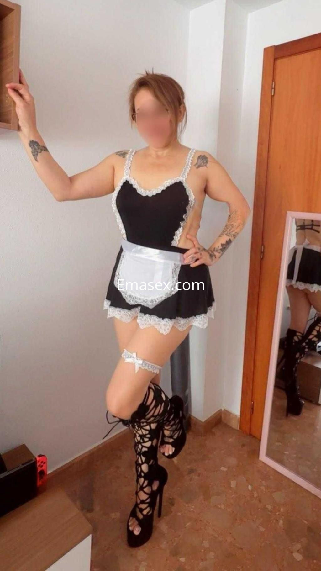 Puta en Alicante viste un disfraz de sirvienta negro con detalles blancos, posando con botas altas y un ambiente moderno.