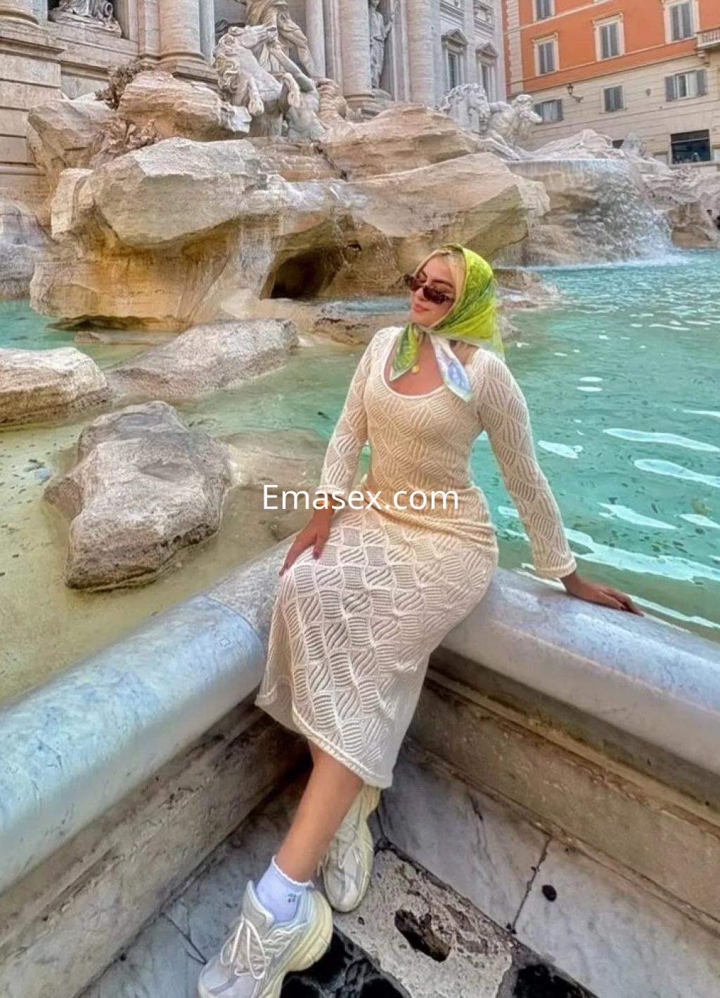 La escort en Oviedo viste un vestido blanco con detalles de encaje y zapatillas, posando junto a una fuente.