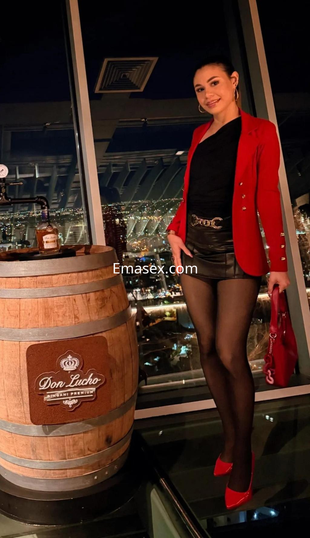 Escort en Málaga, luciendo un blazer rojo, top negro y falda de cuero, posando junto a un barril en un entorno elegante.
