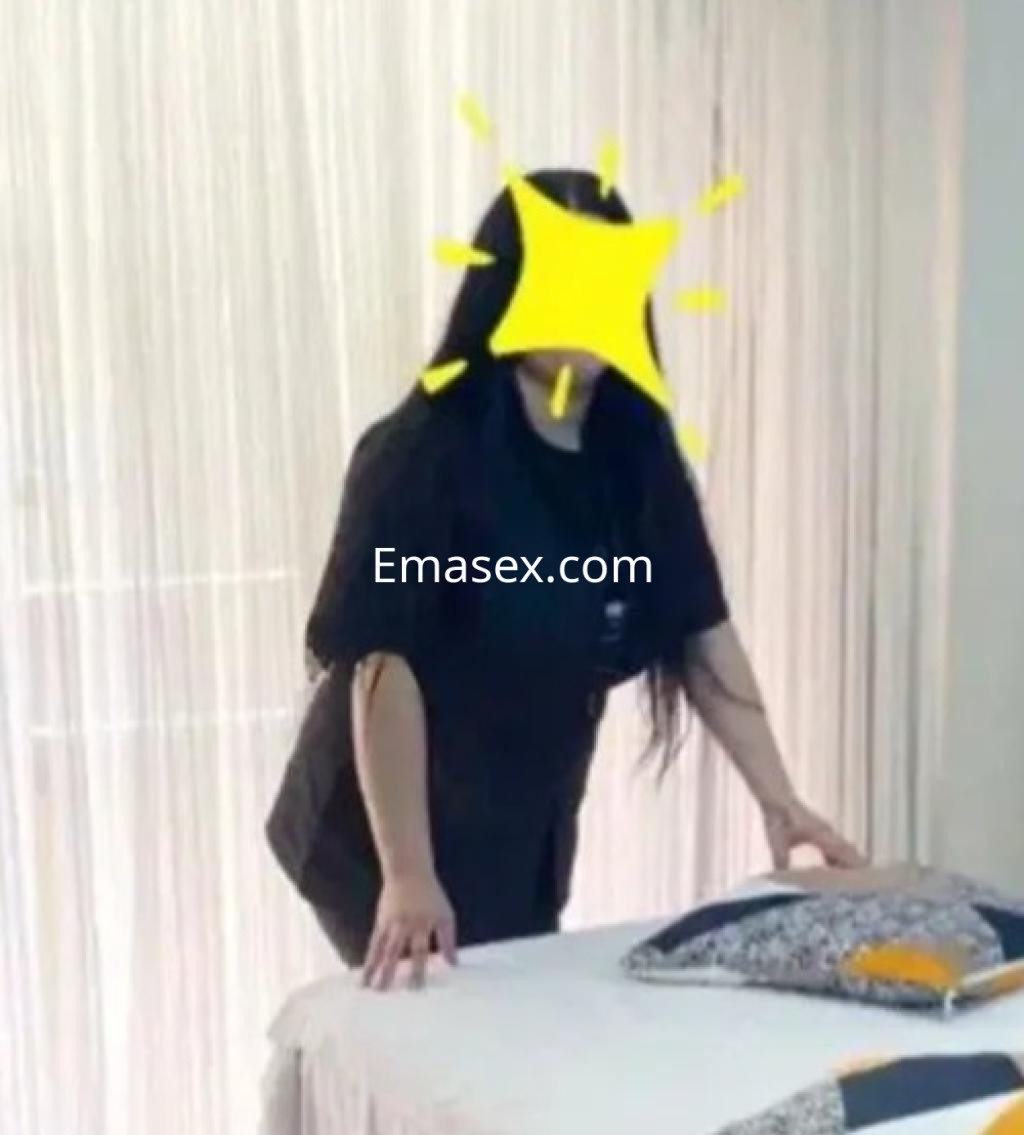 La escort en Oviedo se encuentra de pie junto a una mesa de masaje, vistiendo una camiseta oscura y con cabello largo.