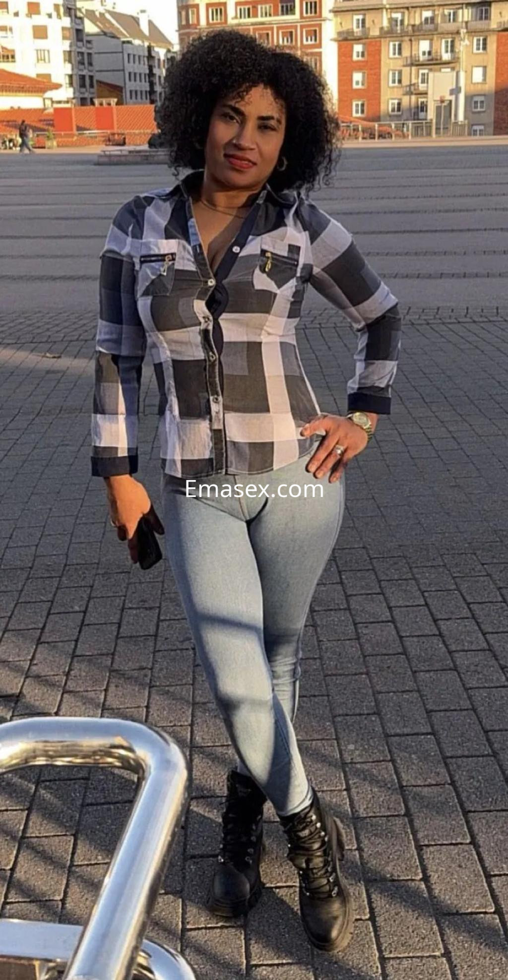 Puta en Oviedo, posando con una camisa de cuadros en blanco y negro y jeans ajustados, luciendo cabello rizado.