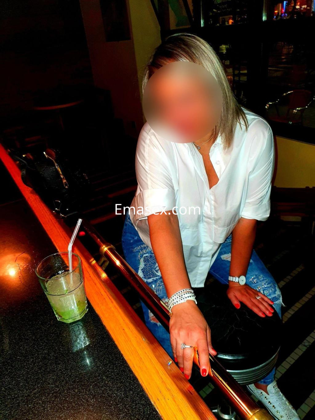 Escort en Valladolid sentada en una barra, luciendo una camisa blanca y jeans, con un cóctel al lado.