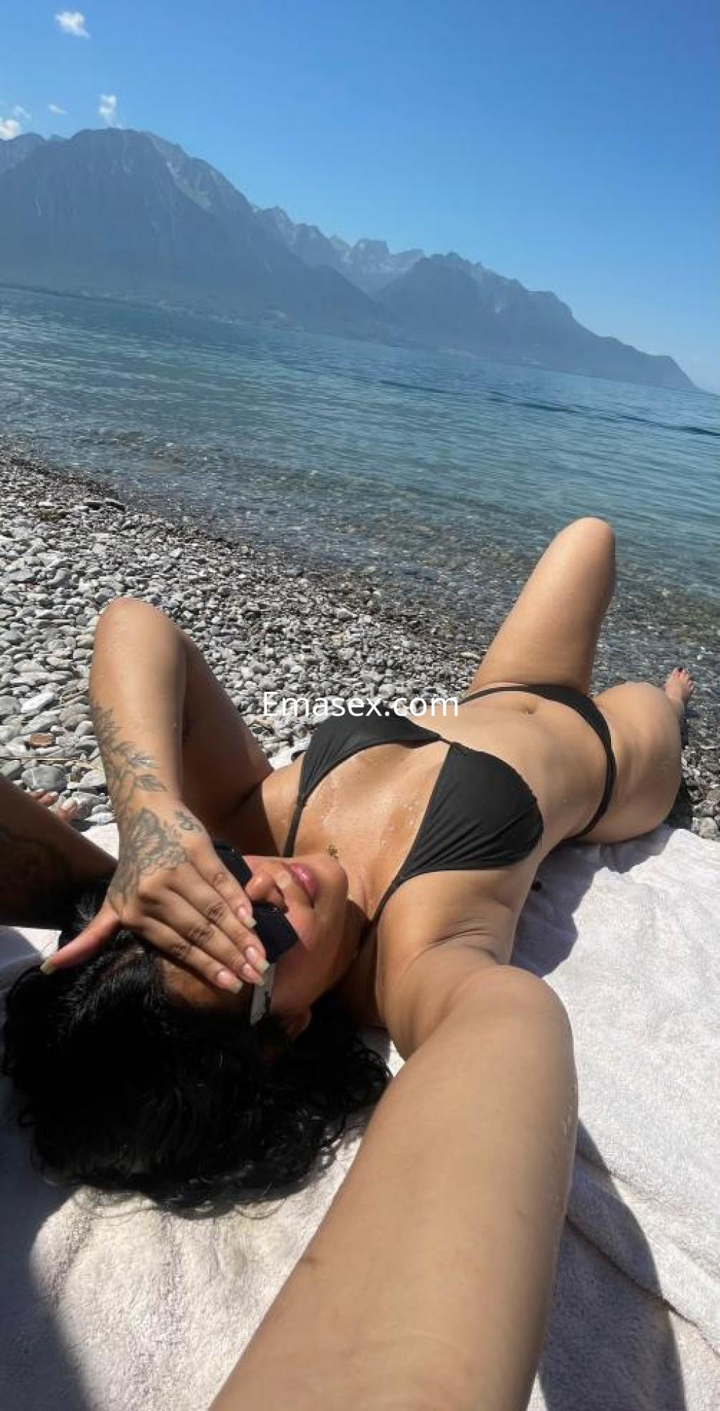 Escort en Madrid, con cabello rizado y oscuro, posando en la playa con un bikini negro y gafas de sol.
