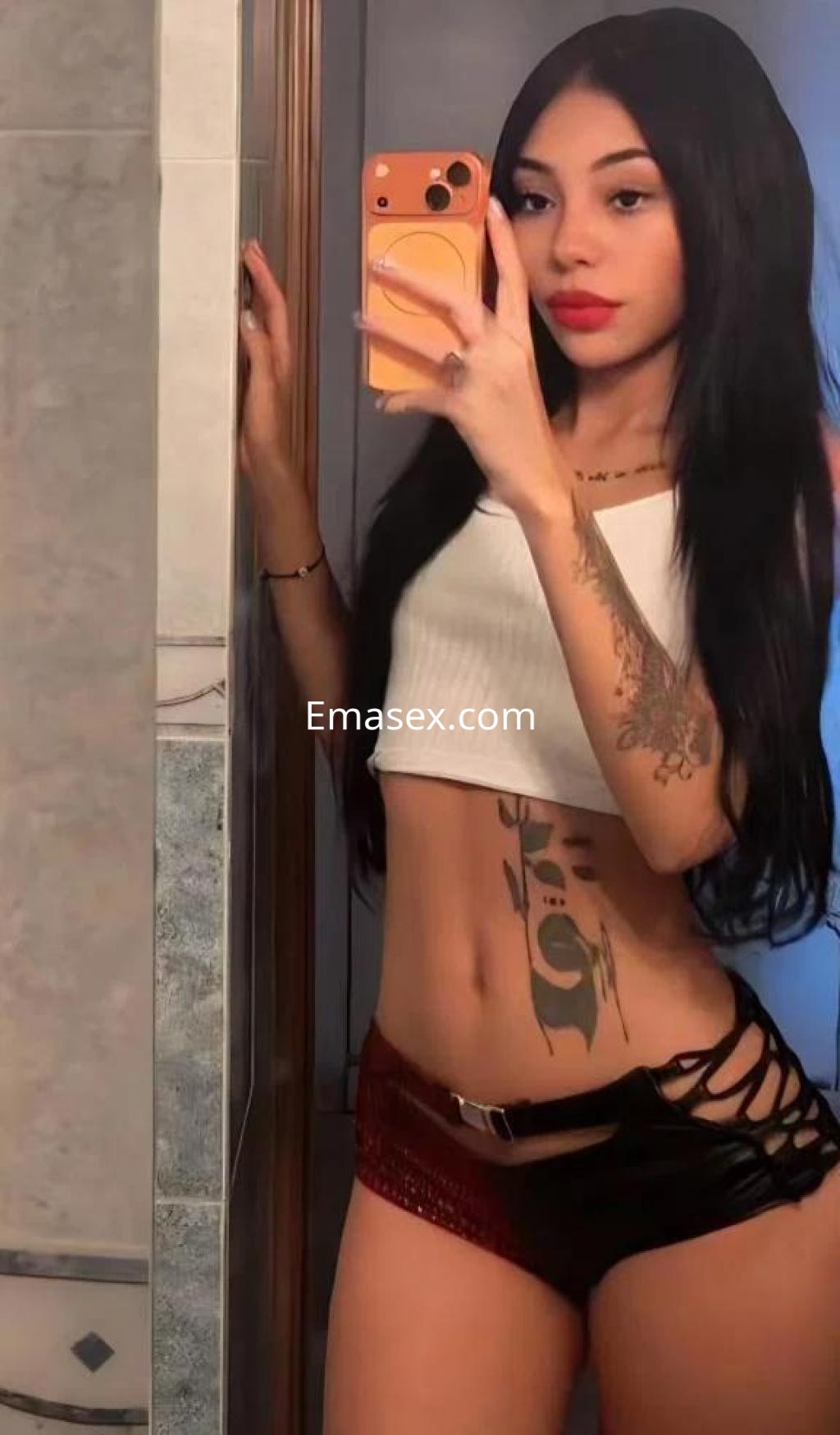Escort en Madrid posando frente al espejo con un top blanco y shorts negros, mostrando un tatuaje en el abdomen.