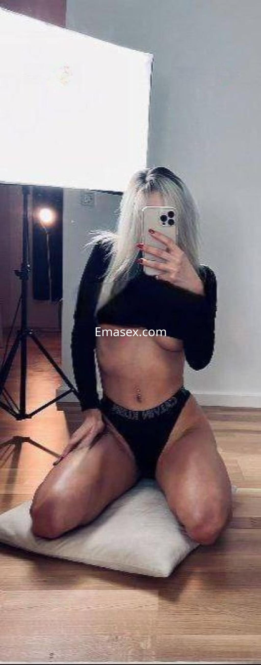 Escort en Málaga posando de rodillas sobre una almohadilla, vistiendo un top negro y braguitas oscuras, con cabello rubio.