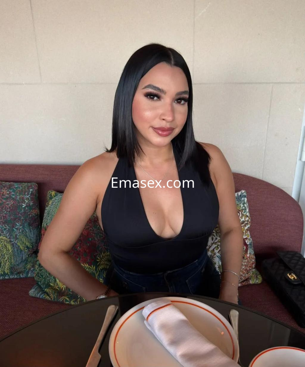Escort en Coruña con cabello lacio y oscuro, viste un top negro y se encuentra sentada en un restaurante elegante.