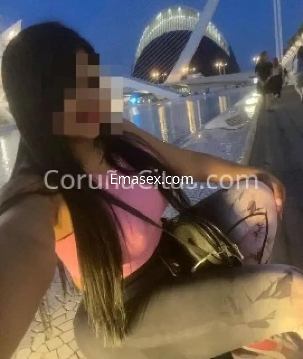 Escort sentada en un muelle de Coruña, vistiendo una blusa rosa y mallas con estampado, con cabello largo.