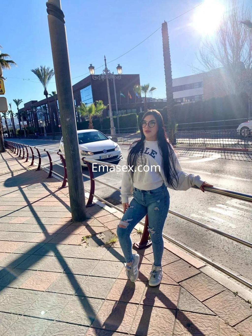 La puta viste un suéter blanco y jeans rasgados, posando con gafas en una soleada calle de Granada.
