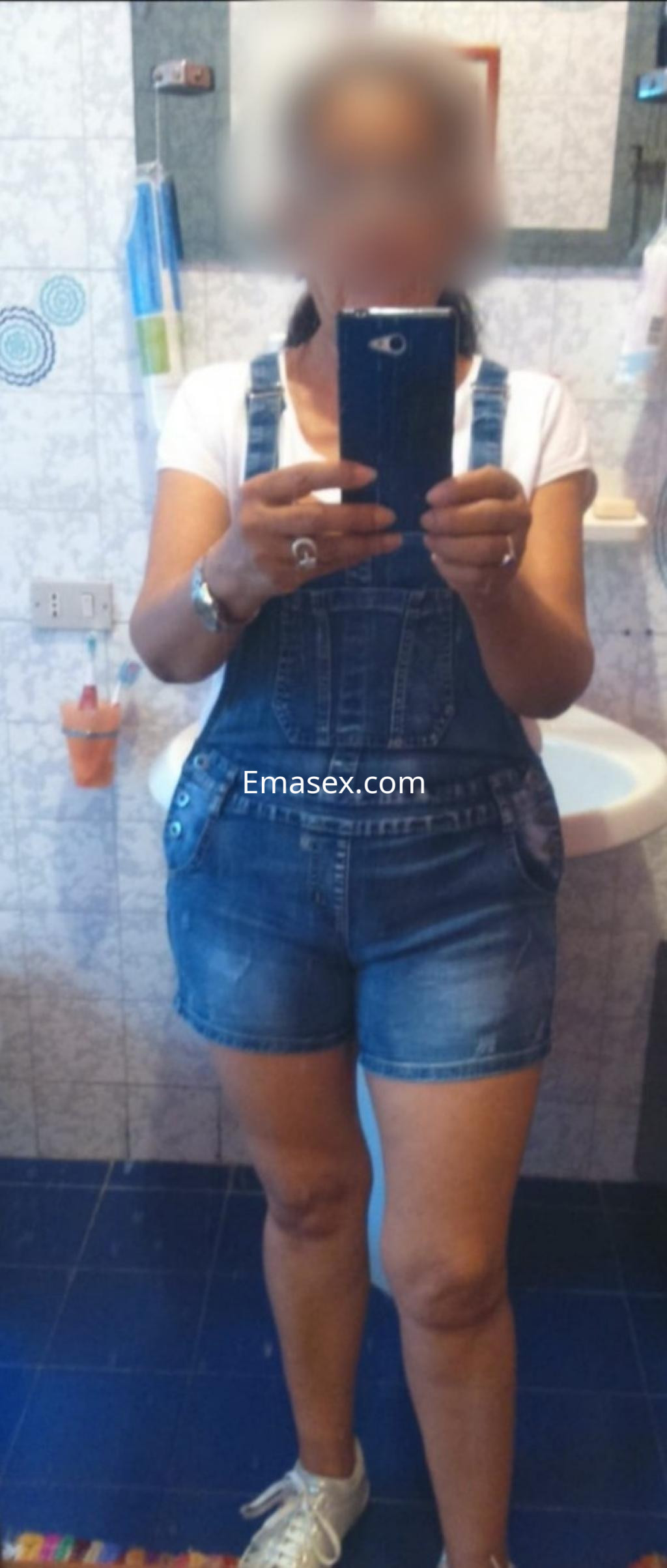 Escort en Santiago con overol de mezclilla y camiseta blanca, posando en un baño con azulejos claros.