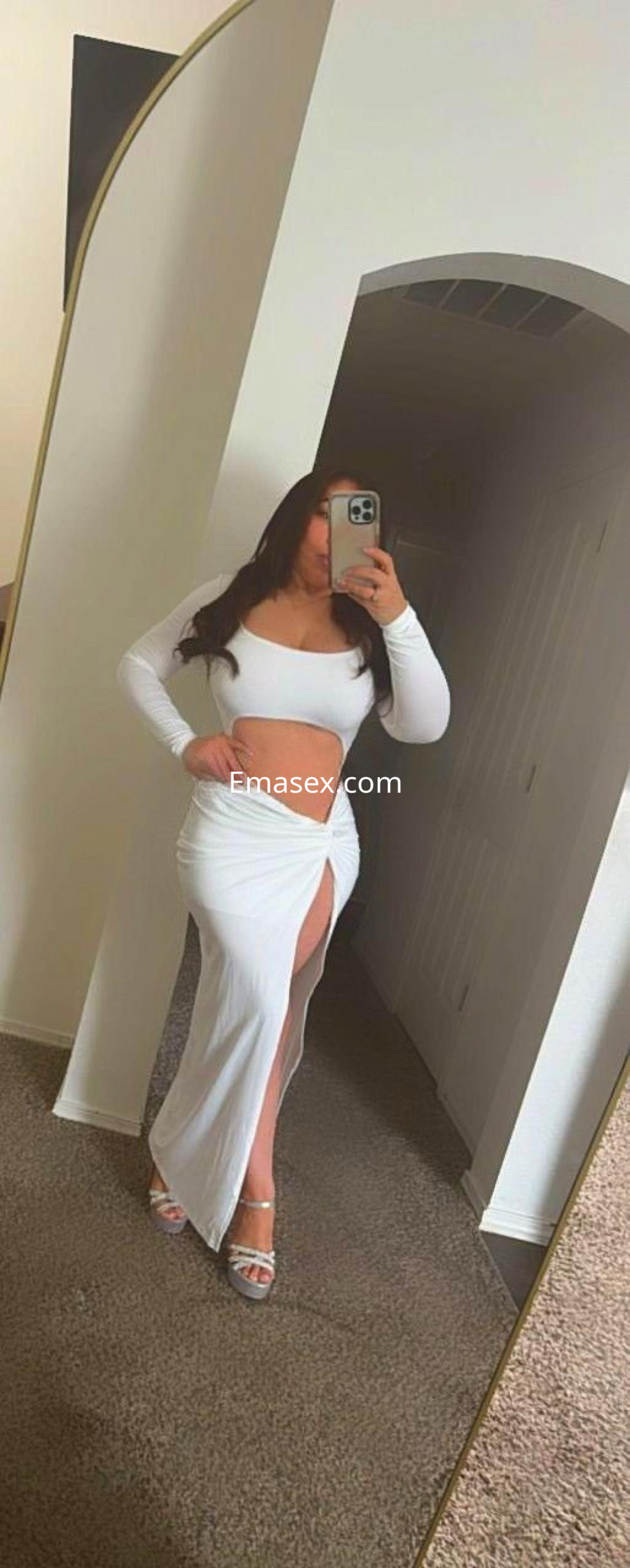 Escort en Madrid luciendo un vestido blanco de dos piezas con abertura lateral y tacones, frente a un espejo.