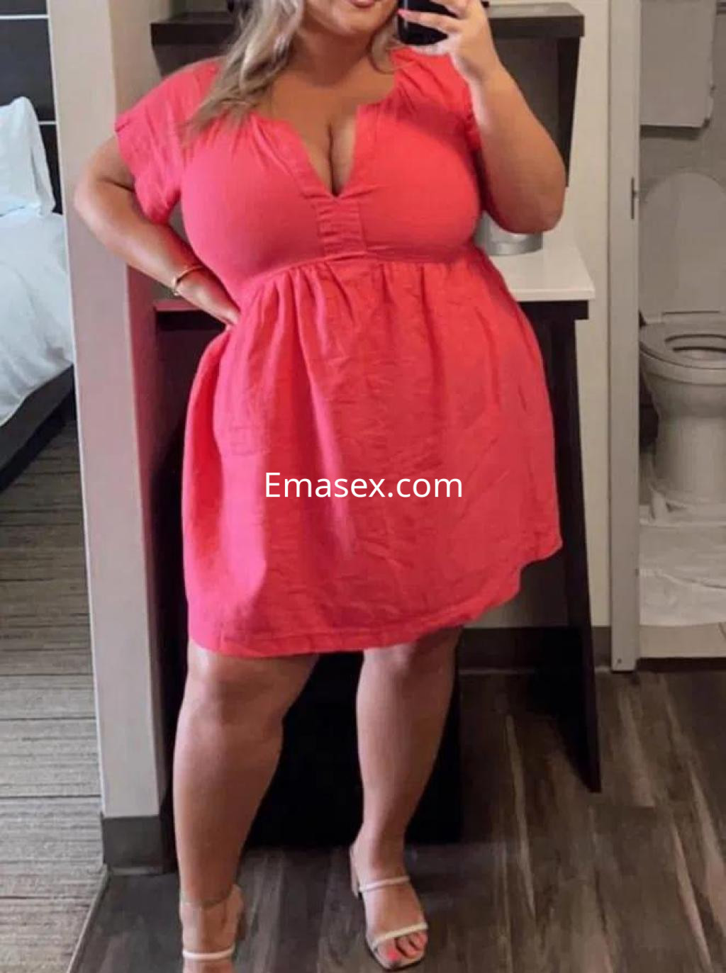 Escort en Ourense con vestido rosa claro, escote pronunciado y sandalias, posando frente a un espejo.