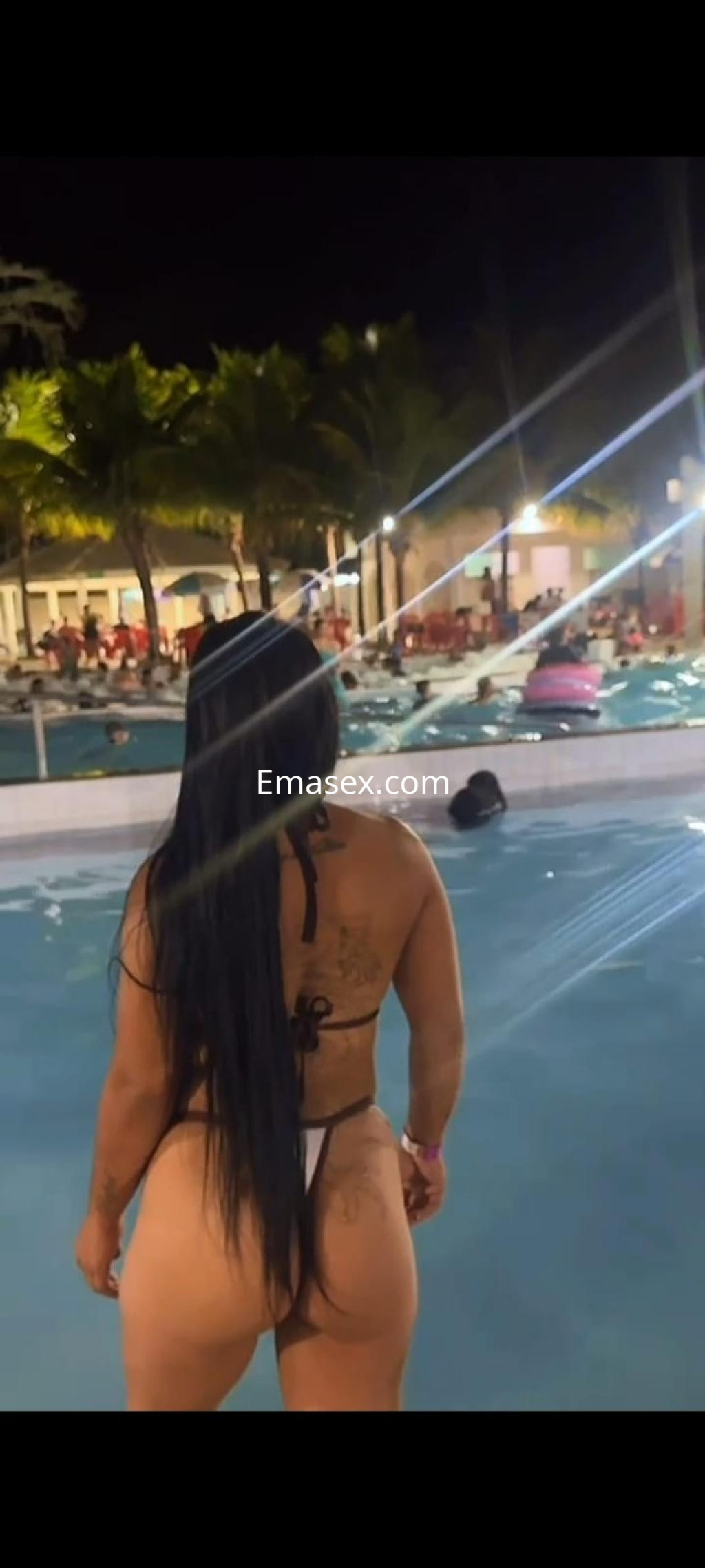 Escort de Ourense de pie junto a una piscina, luciendo un bikini blanco y cabello largo, con un ambiente festivo.