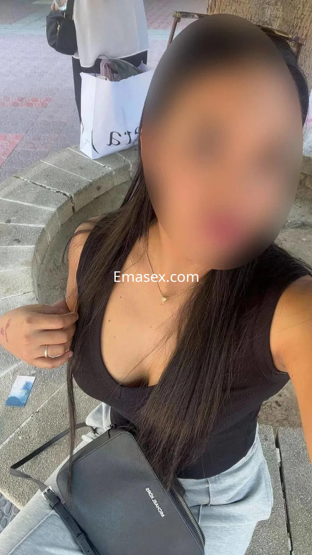 Puta en Zaragoza posando con blusa negra y pantalones claros, sentada en una banca al aire libre.