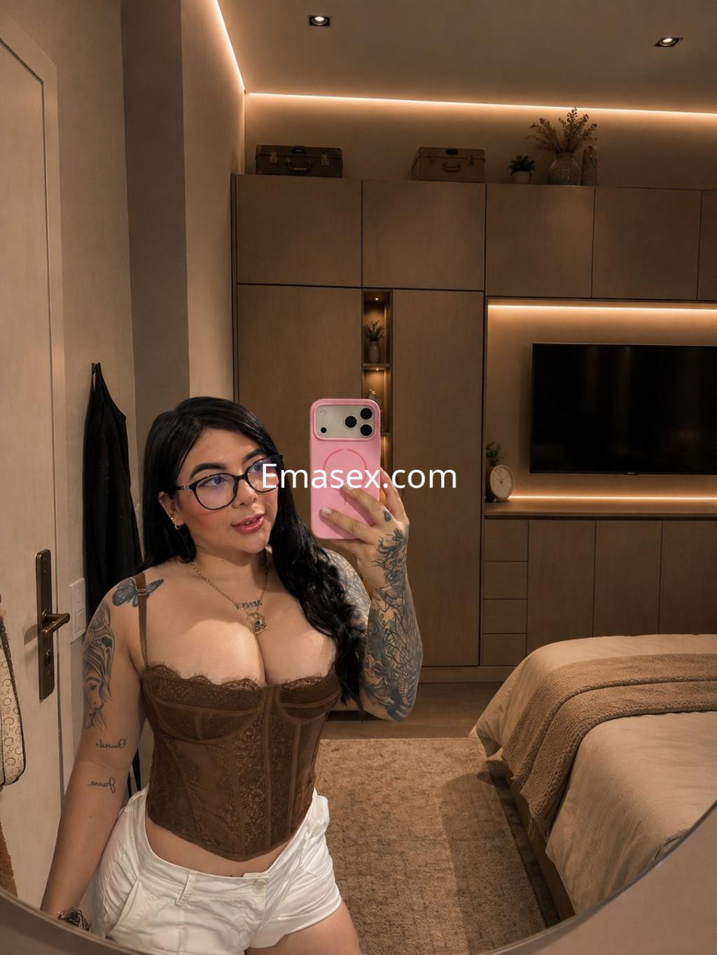 La escort en Bilbao se toma una selfie en un dormitorio, luciendo un corsé marrón y shorts blancos, con cabello largo y lacio.