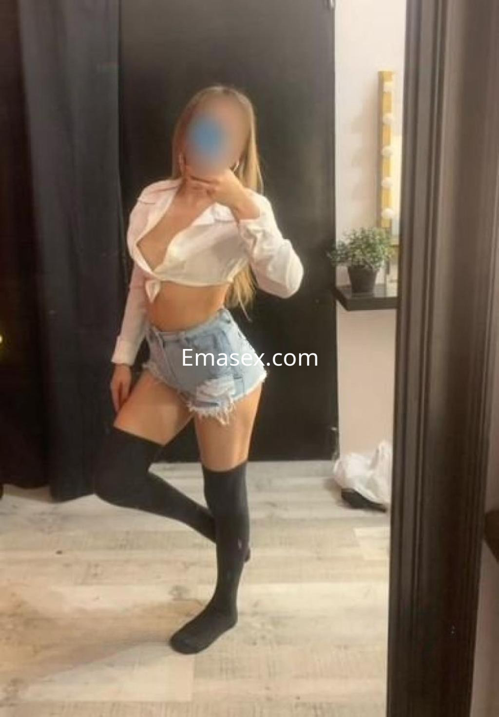 Escort en Barcelona vestida con una blusa blanca, shorts de mezclilla y calcetas negras, posando en un entorno interior.