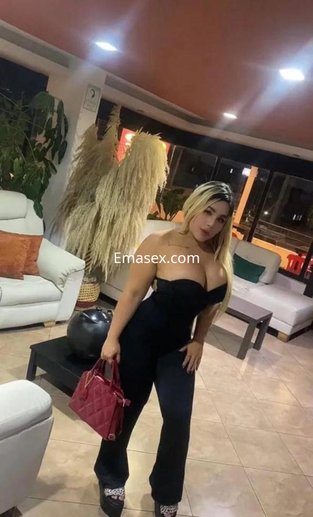 Puta en Valladolid con cabello rubio y lacio, viste un mono negro ajustado y sostiene un bolso rojo en un salón elegante.