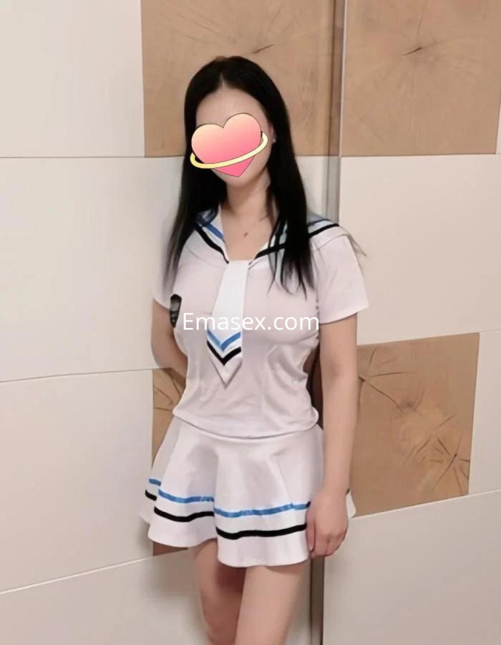 Escort en Ourense con cabello oscuro, vestida con uniforme escolar blanco y azul, posando de pie frente a un fondo claro.