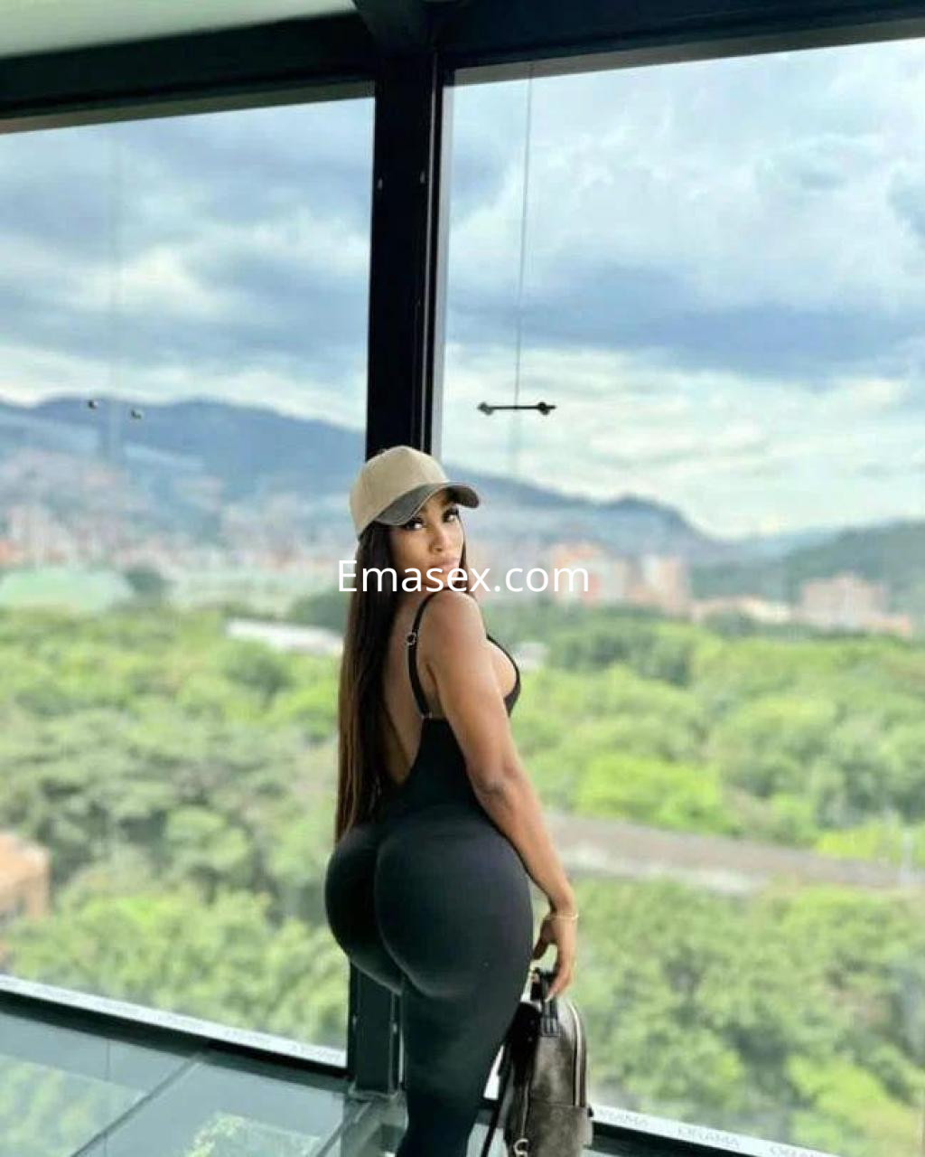 Puta en Girona, de pie frente a una ventana con vista a la ciudad, luciendo un ajustado mono negro y gorra.