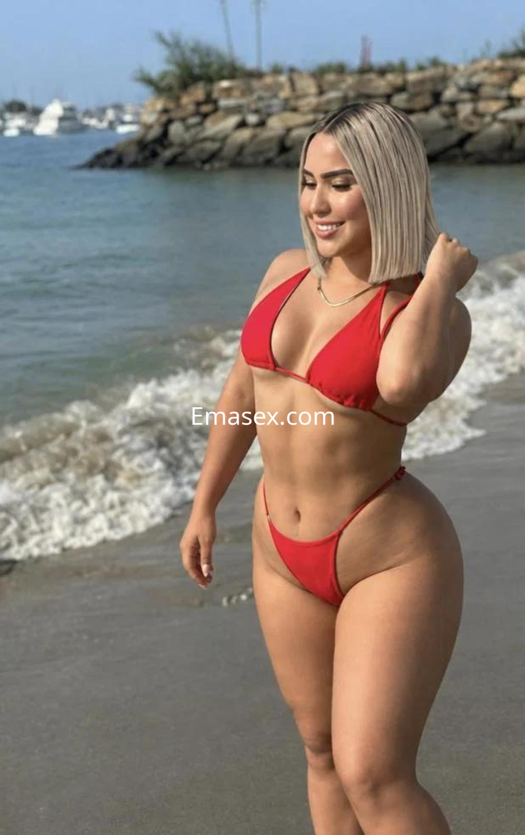 Escort en Las Palmas, con cabello rubio y corto, luciendo un bikini rojo en la playa junto al mar.