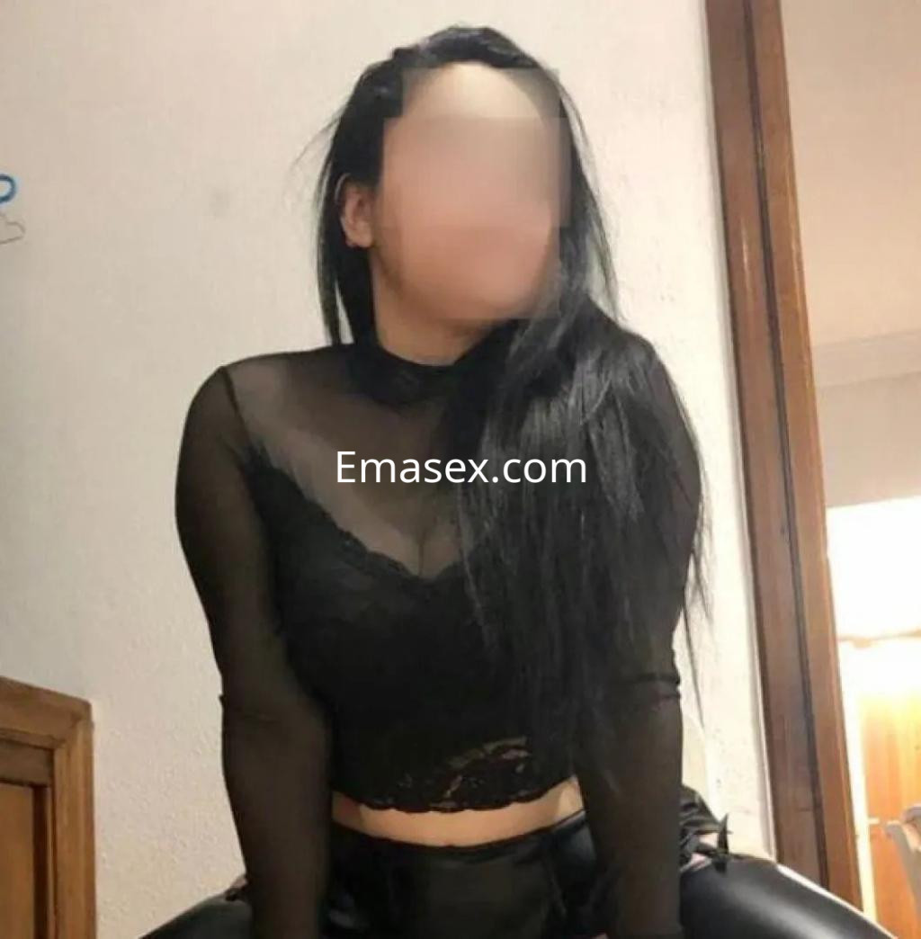 Escort en Córdoba con blusa negra transparente y pantalones ajustados, posando en un entorno interior.