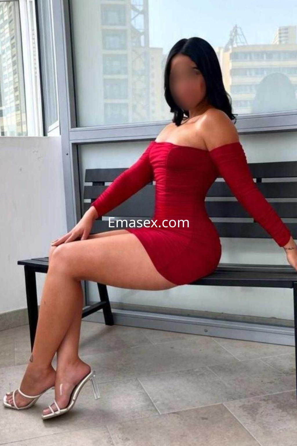 Escort en Alicante, sentada en un banco, viste un vestido rojo ajustado de manga larga y tacones plateados.