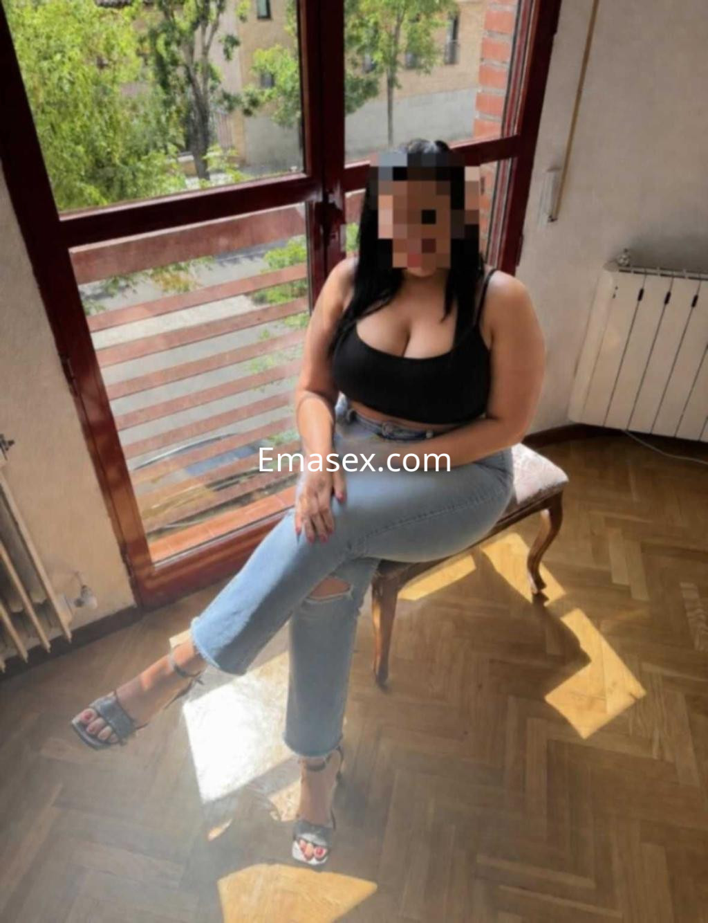 Escort en Toledo, sentada en una silla con piernas cruzadas, viste top negro y jeans claros, con sandalias.