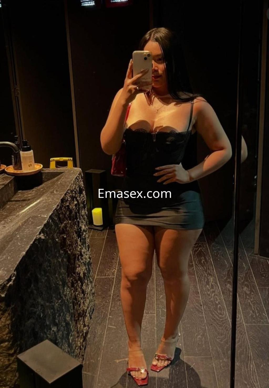 Puta con vestido negro ajustado y tacones en un ambiente oscuro, posando frente a un espejo en Santiago.