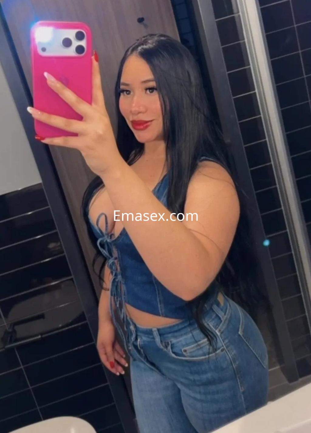 Escort en Alicante con cabello largo y lacio, viste un top de mezclilla y jeans ajustados, posando en un espejo.