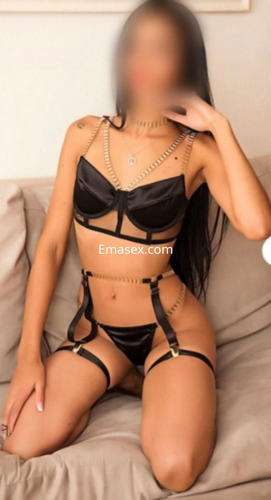 Escort en Madrid posando de rodillas sobre un sofá, vistiendo lencería negra con detalles dorados y cabello largo.