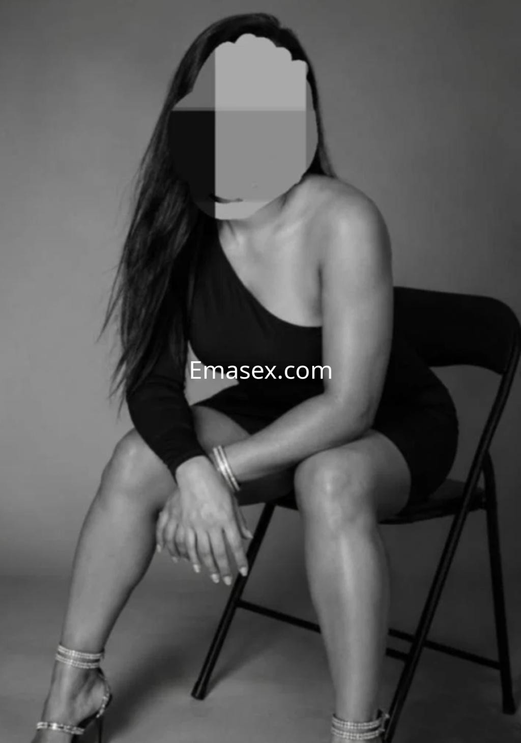 Escort sentada en una silla con un vestido negro de un solo hombro, en un fondo gris en Logroño.