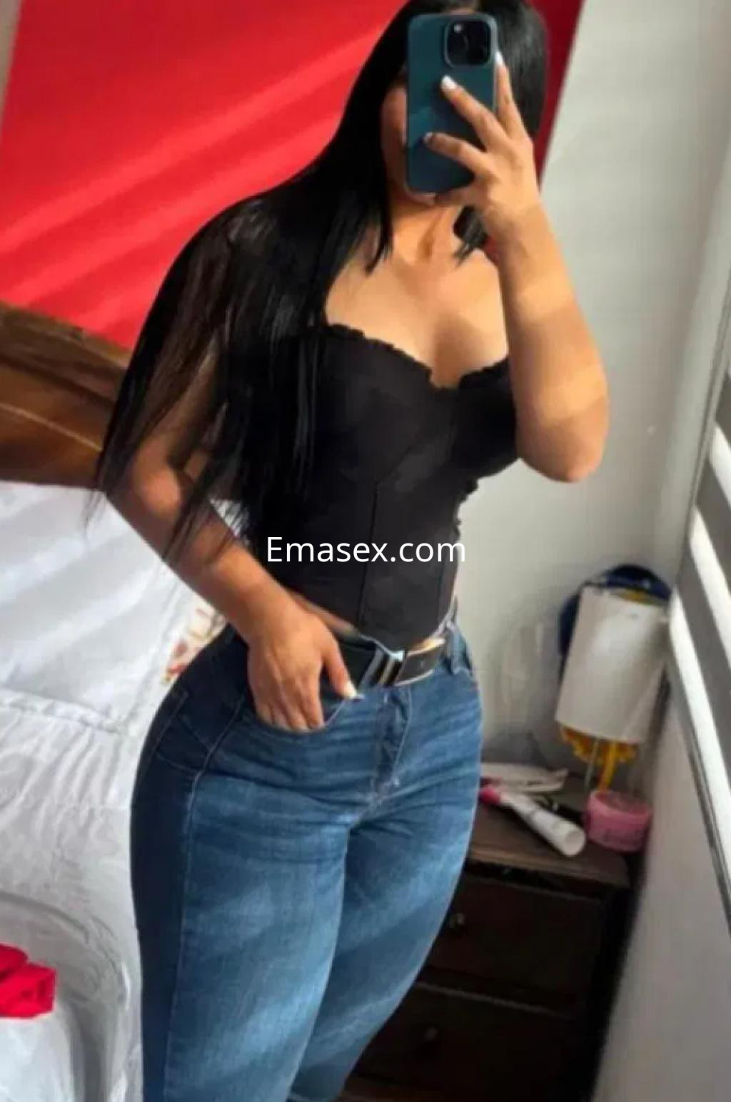 Escort en Pamplona luciendo un top negro ajustado y jeans, con cabello largo y lacio, posando en un dormitorio.