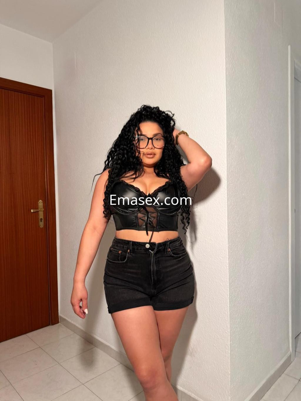 Escort en Málaga con pelo rizado y oscuro, viste un top de cuero negro y shorts de mezclilla, en una pose segura.