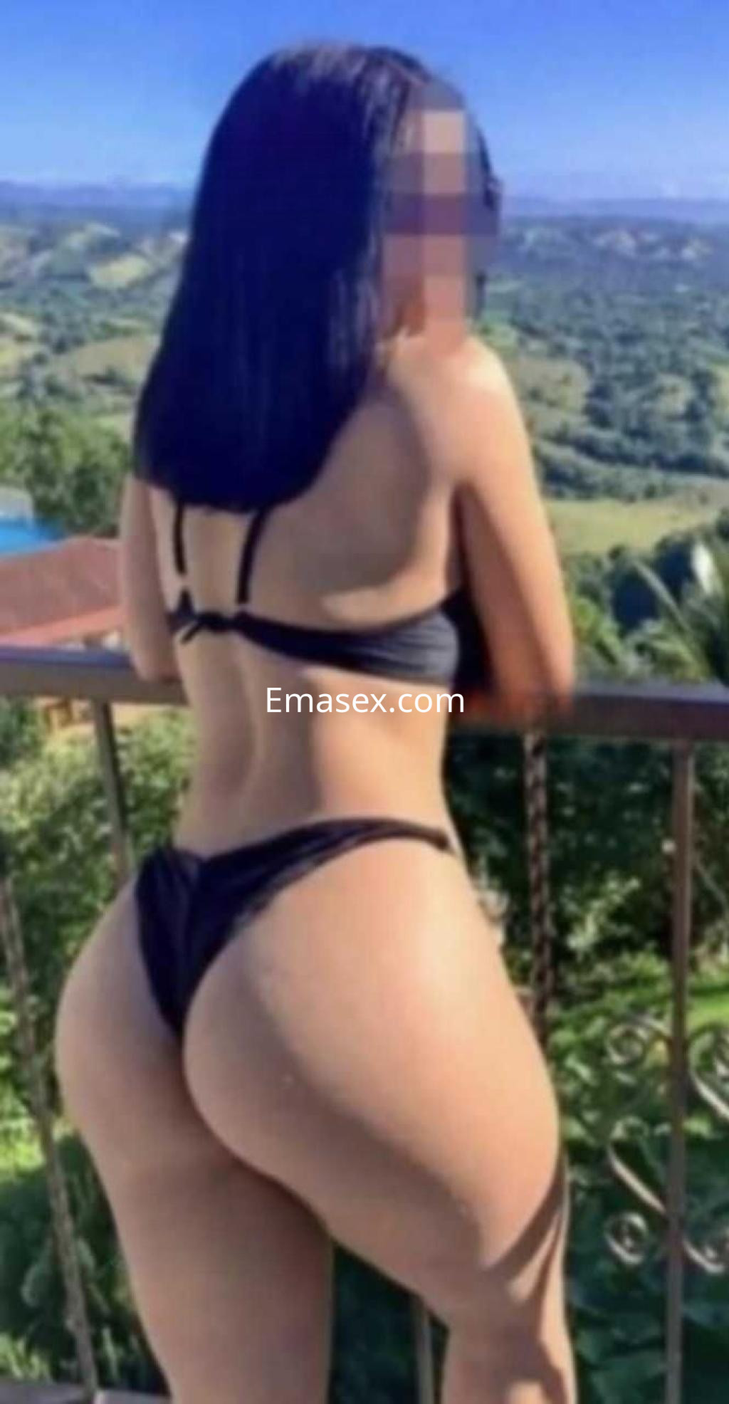 Puta posando de espaldas en Tarragona, con un bikini negro y el cabello lacio, frente a un paisaje verde.