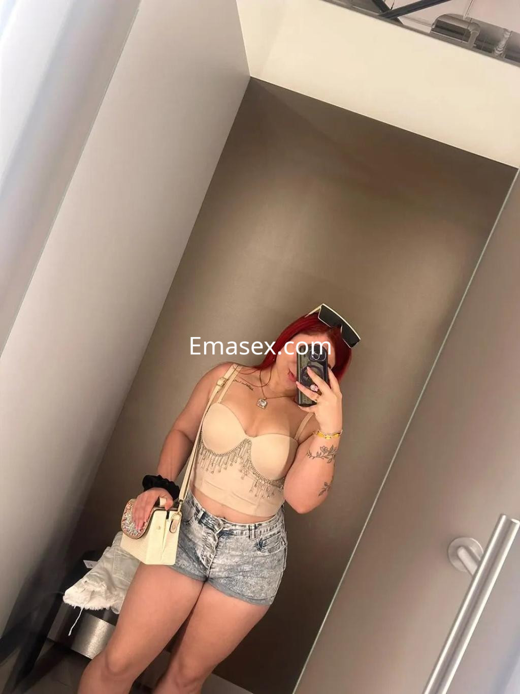 Puta en Vigo, frente a un espejo, con un sujetador con detalles brillantes y shorts de mezclilla, sosteniendo un bolso.