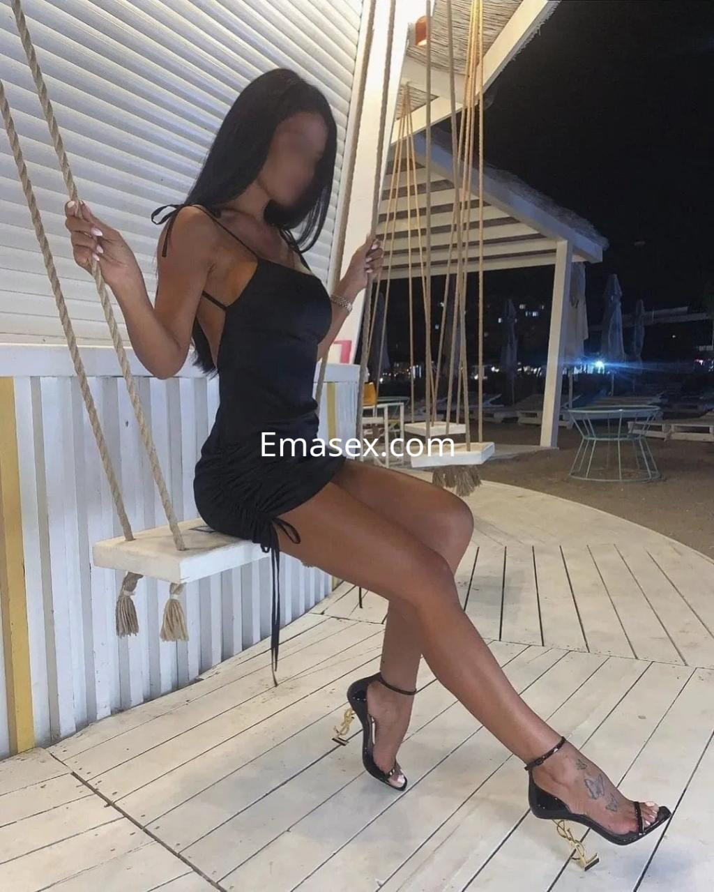 Escort en Alicante sentada en un columpio, luciendo un vestido negro ajustado y tacones elegantes.