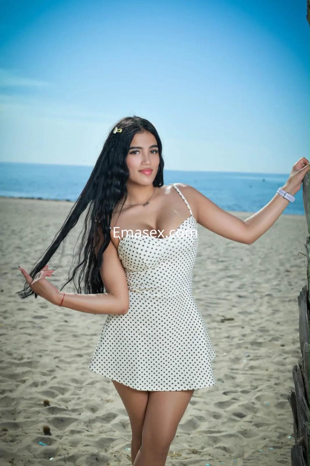 Escort en Alicante posando en la playa con vestido blanco de lunares y cabello largo, sonriente y relajada.