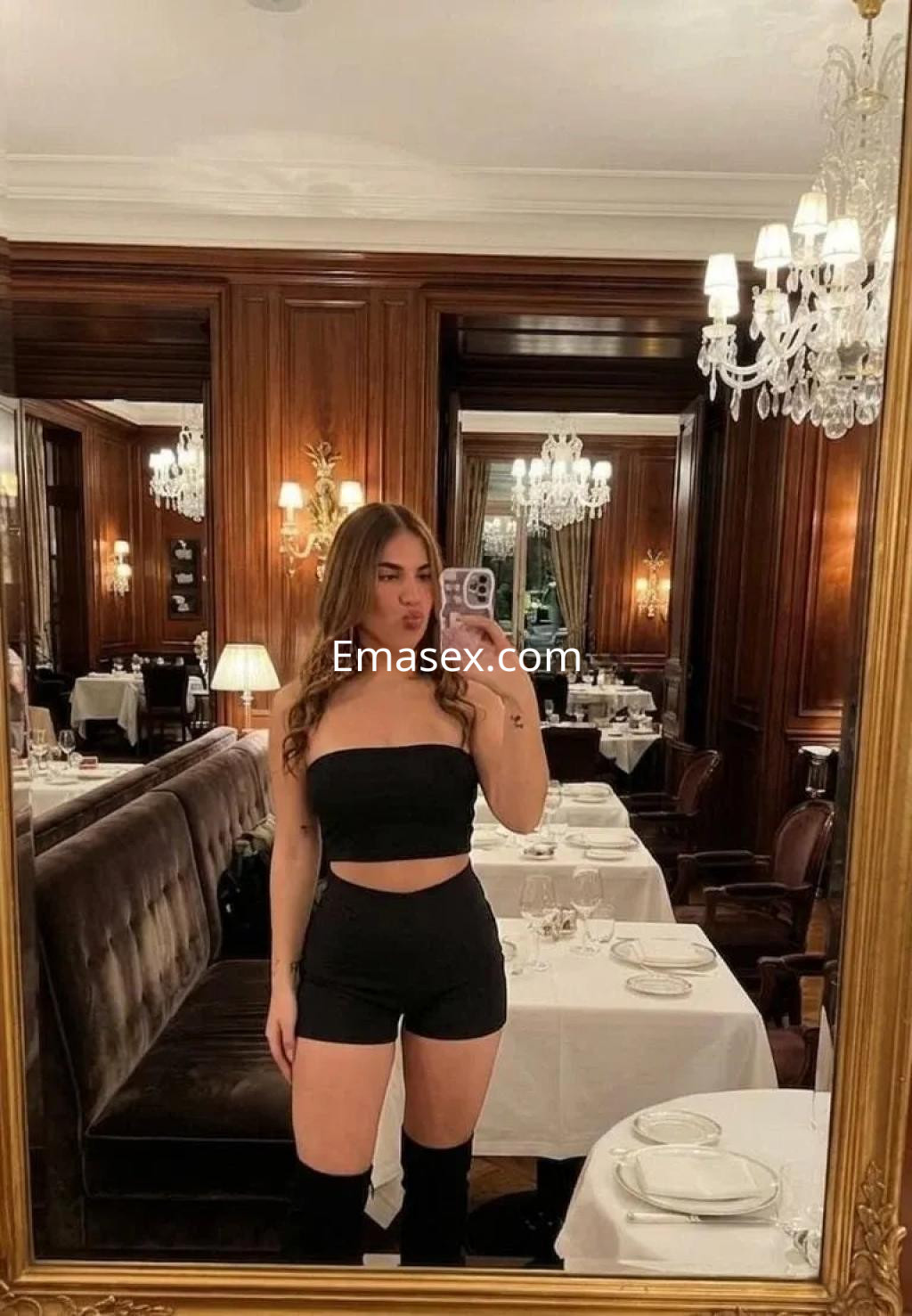 Escort en Alicante con cabello largo y rizado, luciendo un conjunto negro y posando en un elegante restaurante.