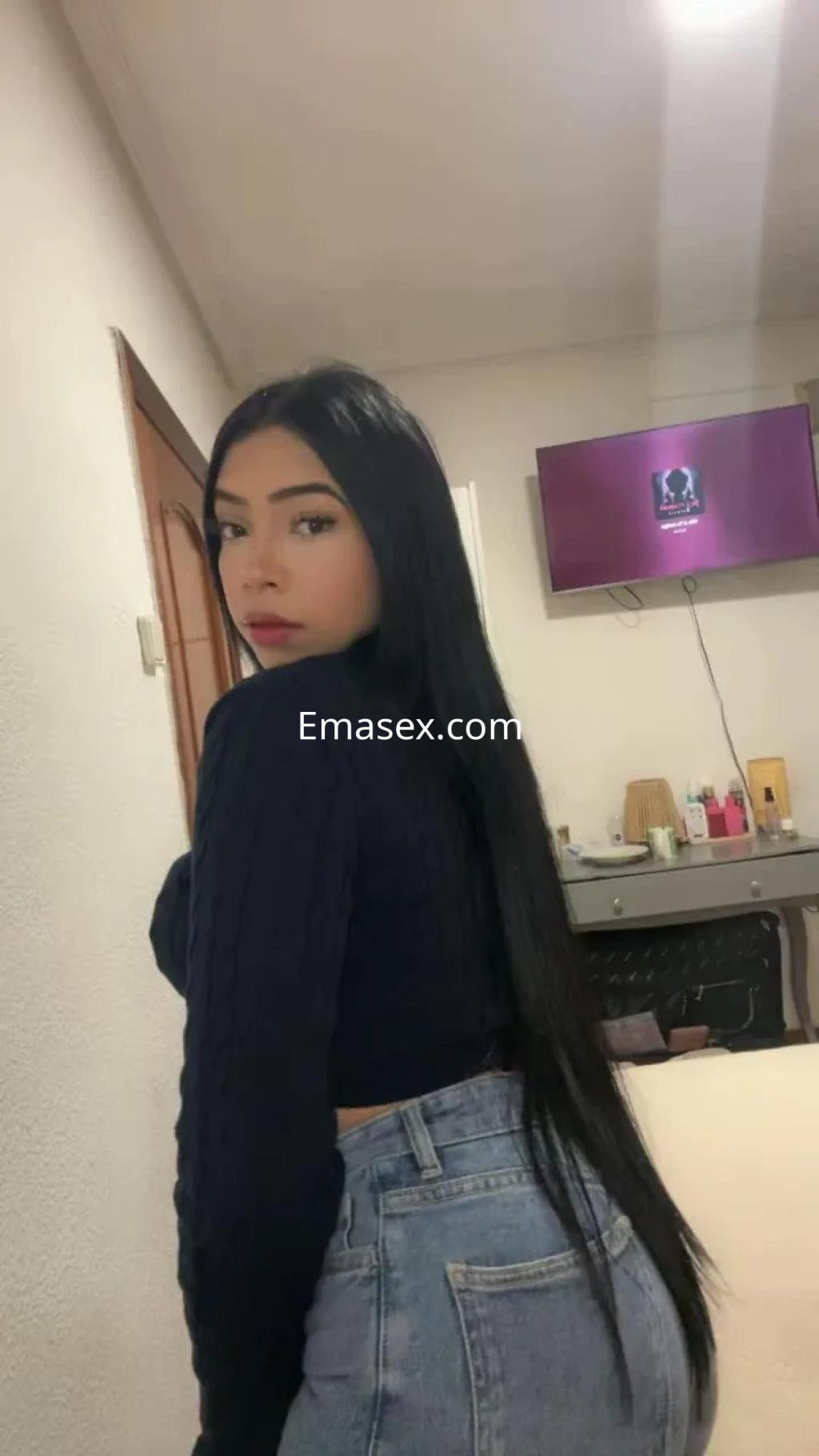 Escort en Coruña con cabello largo y lacio, viste un suéter azul y jeans ajustados, posando en un ambiente interior.
