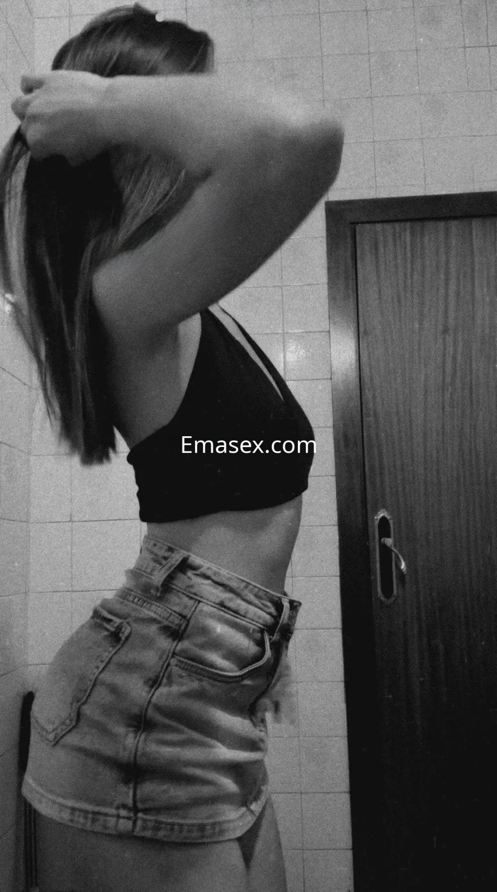 Escort en Córdoba con cabello lacio y oscuro, viste un top negro y shorts de mezclilla, posando de perfil.