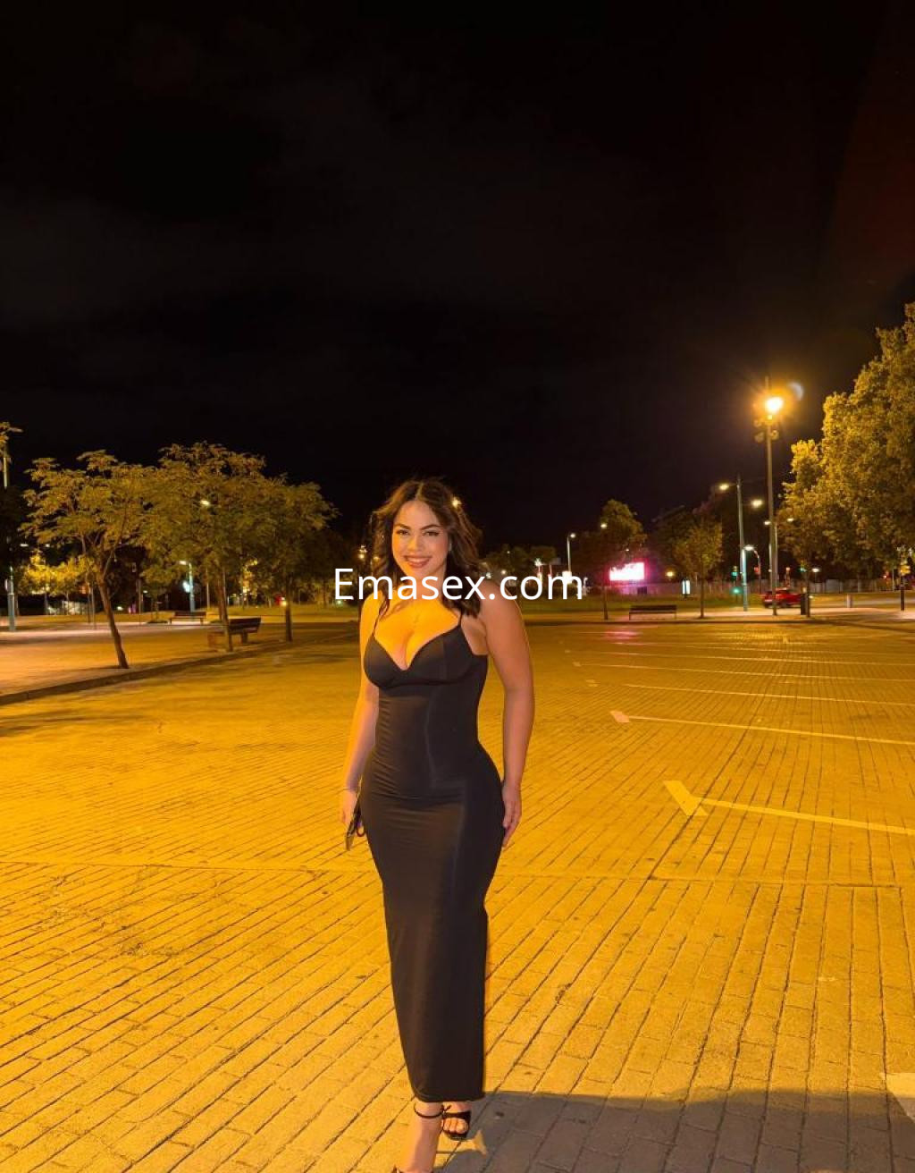Puta de Zaragoza posando al aire libre con un vestido negro ajustado, en un entorno iluminado por faroles.