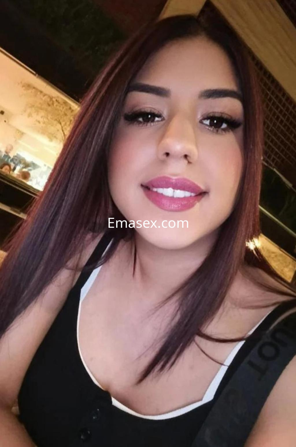 Escort en Huelva con cabello corto y lacio, vistiendo una blusa negra, sonriendo en un ambiente iluminado.