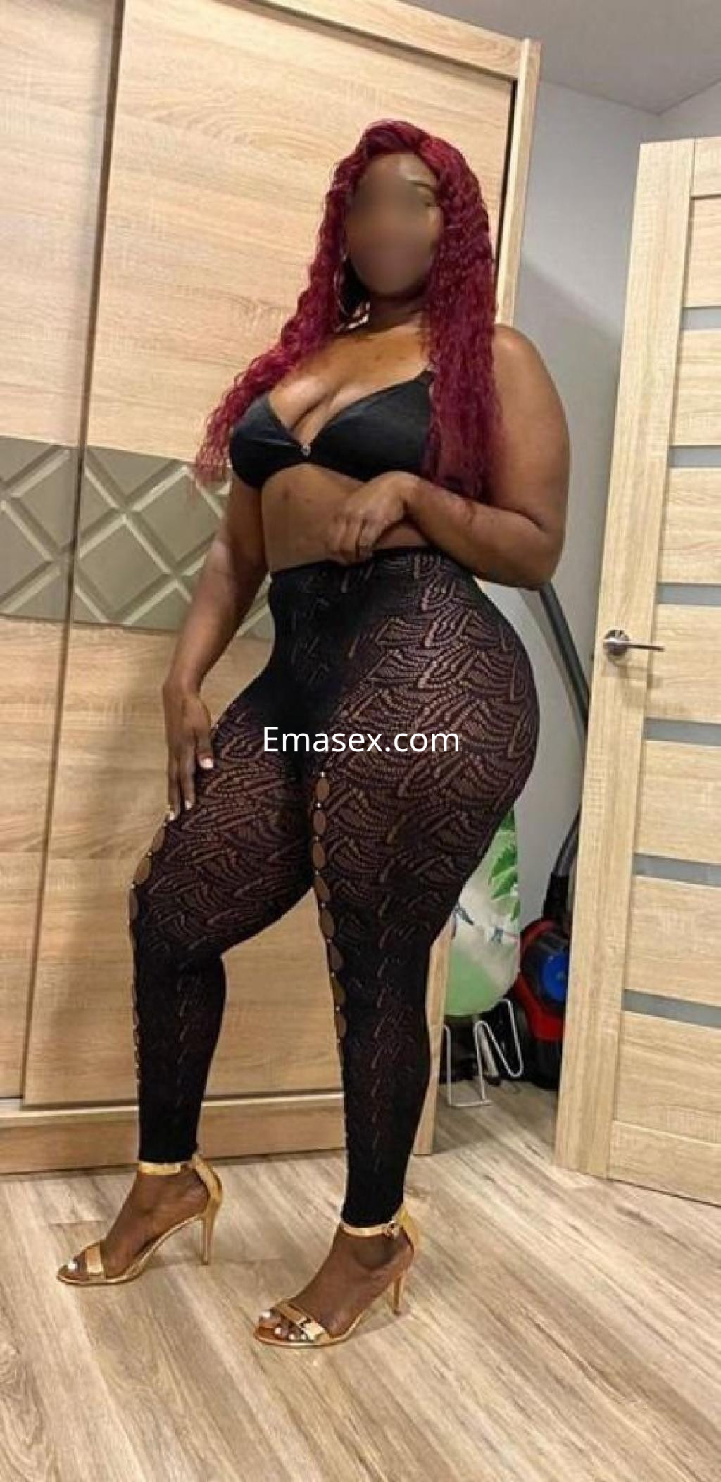 Escort posando en Huelva, con cabello rizado y rojo, vistiendo un top negro y leggings de encaje, sandalias doradas.